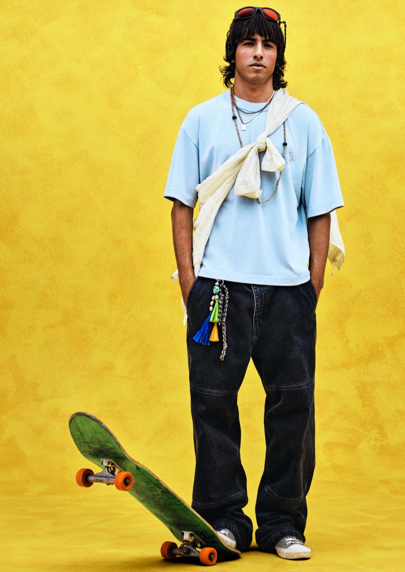 A young model stands against a yellow backdrop in a baby blue tee with a cream shirt knotted over the shoulders, dark wide-leg cargo trousers, and white sneakers, with a green skateboard at his feet