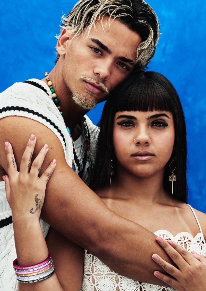 Close portrait of Arthur and Geovanna against a blue backdrop, with Arthur in a white knit tank and bright beaded necklaces resting his head against Geovanna, who wears a white crochet top and a sleek dark fringe