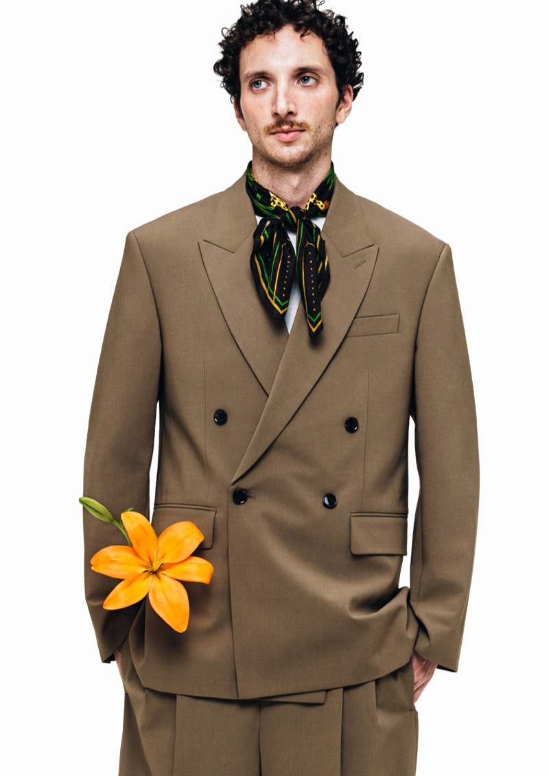 Dadá stands in an oversized tan double-breasted suit and a green and yellow printed silk neckerchief, holding an orange lily
