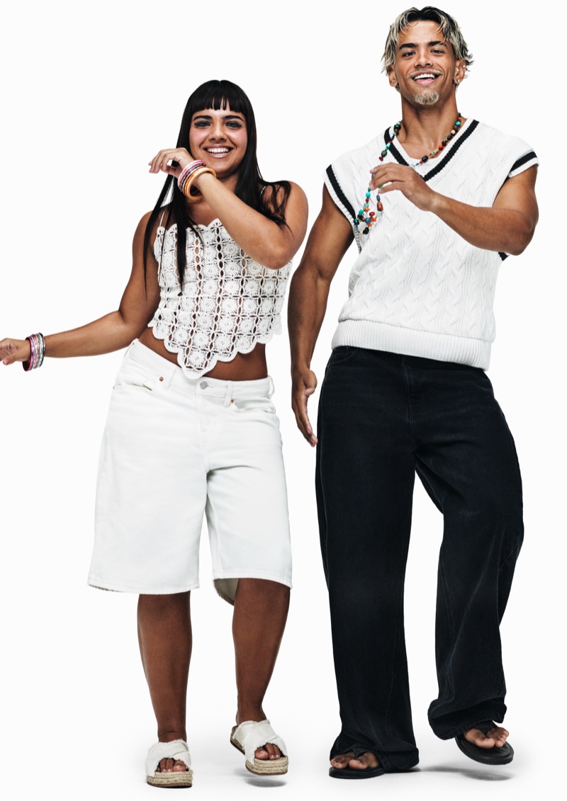 Geovanna in a white crochet crop top and white denim long shorts beside Arthur in a white cricket sweater vest with black trim and dark wide-leg trousers, both moving against a white backdrop