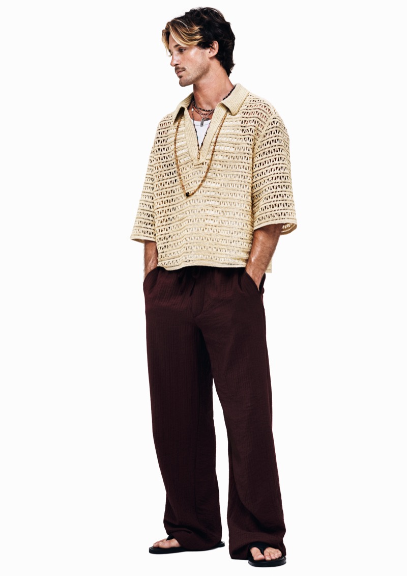 Luca stands in profile in a cream open-knit short-sleeve polo over a white tee with wide-leg brown linen trousers, black flip-flops, and stacked beaded necklaces