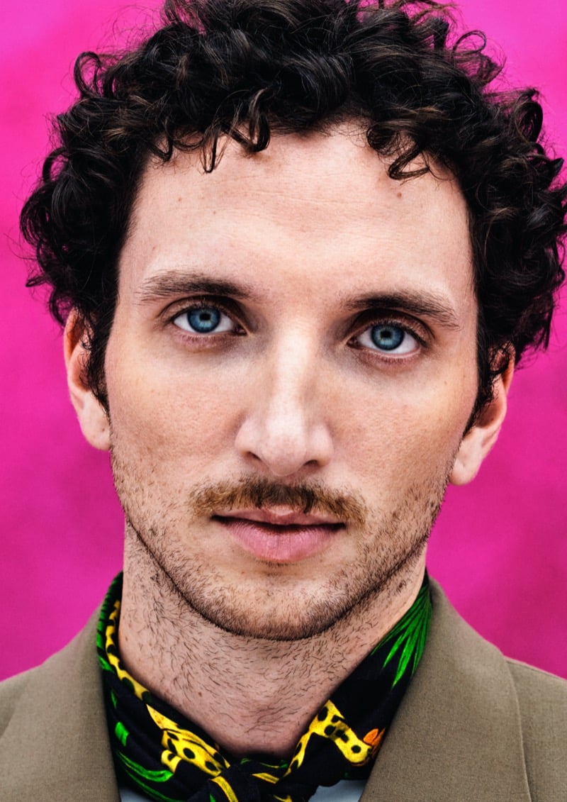 Close portrait of Dadá against a hot pink backdrop with curly dark hair, light eyes, and a green and yellow printed silk neckerchief over a tan jacket