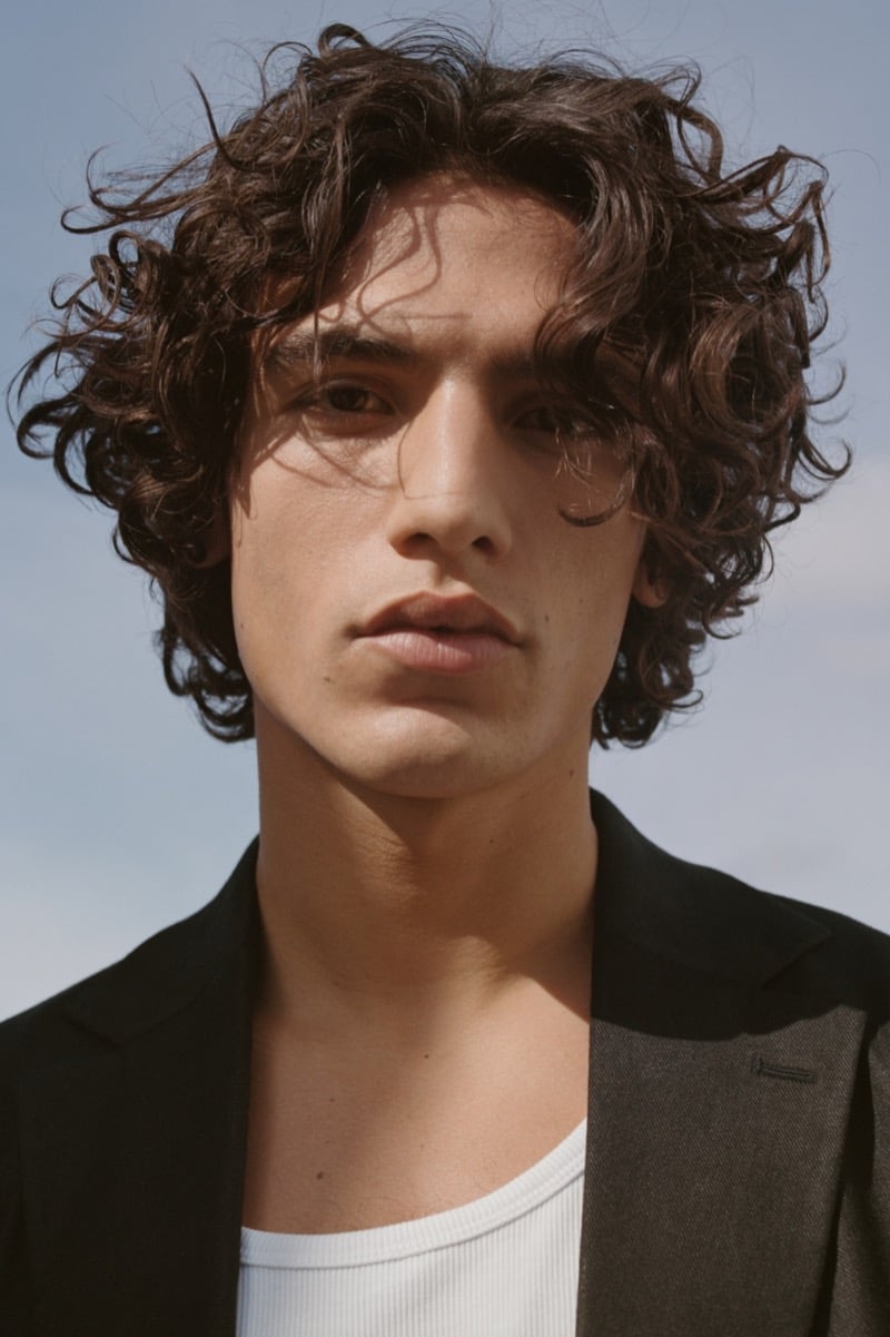 Close-up portrait of Morgan with curly dark hair against an open sky, wearing a dark single-button tailored jacket open over a white ribbed tank top
