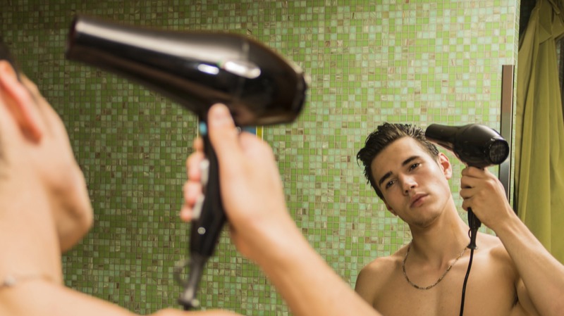 young man with short dark hair stands shirtless in front of a green mosaic-tile bathroom wall, lifting a black blow dryer toward the side of his head as he watches himself in the mirror