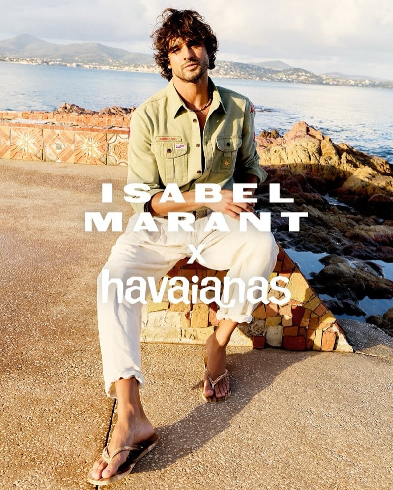Marlon Teixeira sits on a mosaic-tiled stone seawall with the Mediterranean and distant hills behind him, wearing an olive-green military overshirt with flap pockets and embroidered patches over cuffed white linen trousers, sand tie-dye Isabel Marant x Havaianas thong sandals on his feet, dark curly hair tousled in the late afternoon sun