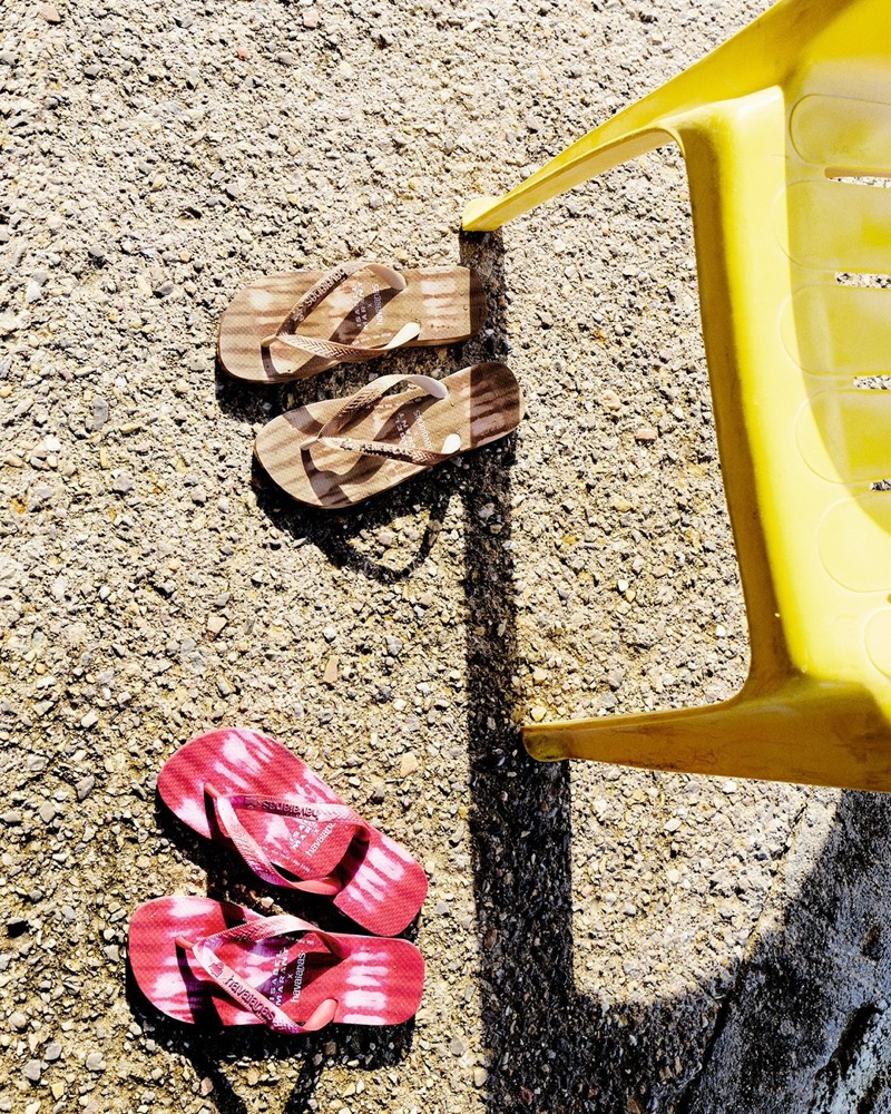 Overhead view of two pairs of Isabel Marant x Havaianas thong sandals laid out on sun-bleached sandy concrete beside a yellow monobloc chair casting a sharp shadow