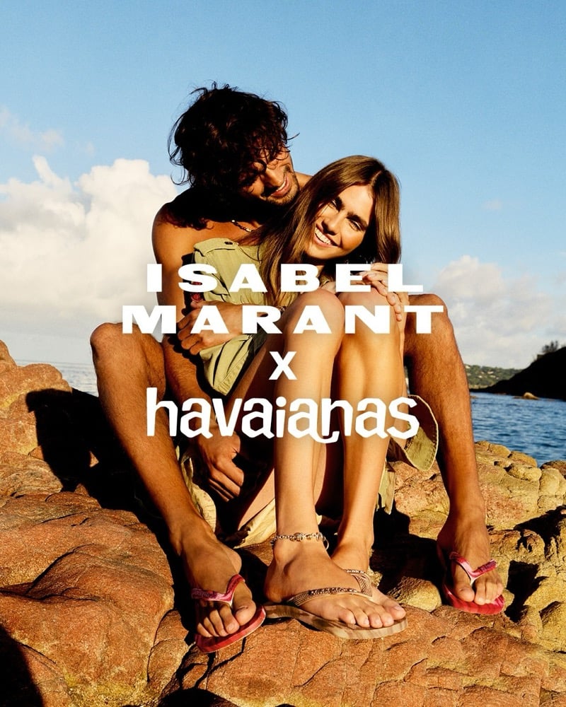 Marlon Teixeira and Mia Armstrong sit close together on weathered red rocks at the edge of the sea under a soft blue sky, his arms loosely around her shoulders, both barefoot in the campaign's Isabel Marant x Havaianas thong sandals — red tie-dye on him and sand tie-dye on her