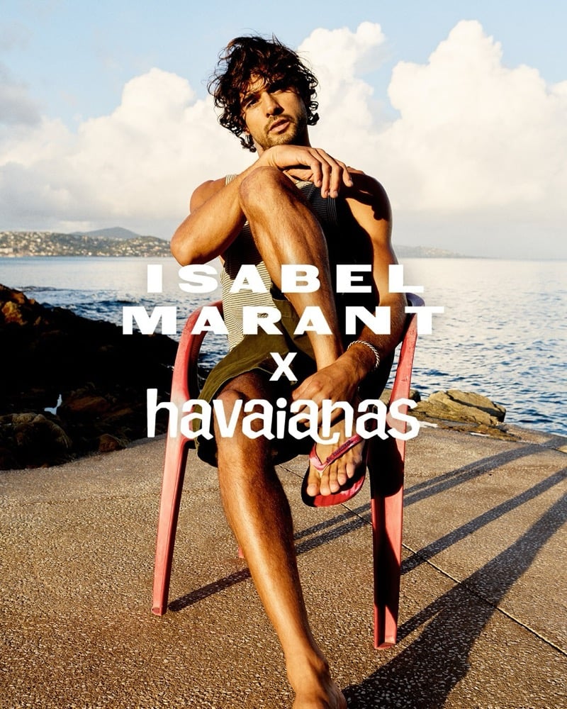Marlon Teixeira sits low in a coral-red monobloc chair on a stone jetty above the sea, one leg crossed over the other, wearing a dark olive ribbed tank top and matching khaki olive shorts with the tie-dye Isabel Marant x Havaianas thong sandals on his feet, dark curly hair and beard catching the sunset light
