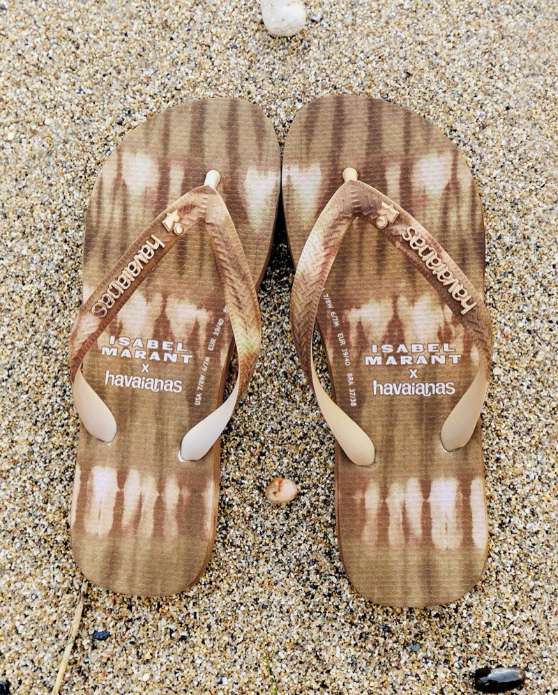 Close-up of the sand and brown tie-dye Isabel Marant x Havaianas thong sandals placed on pale beach sand with small pebbles around them