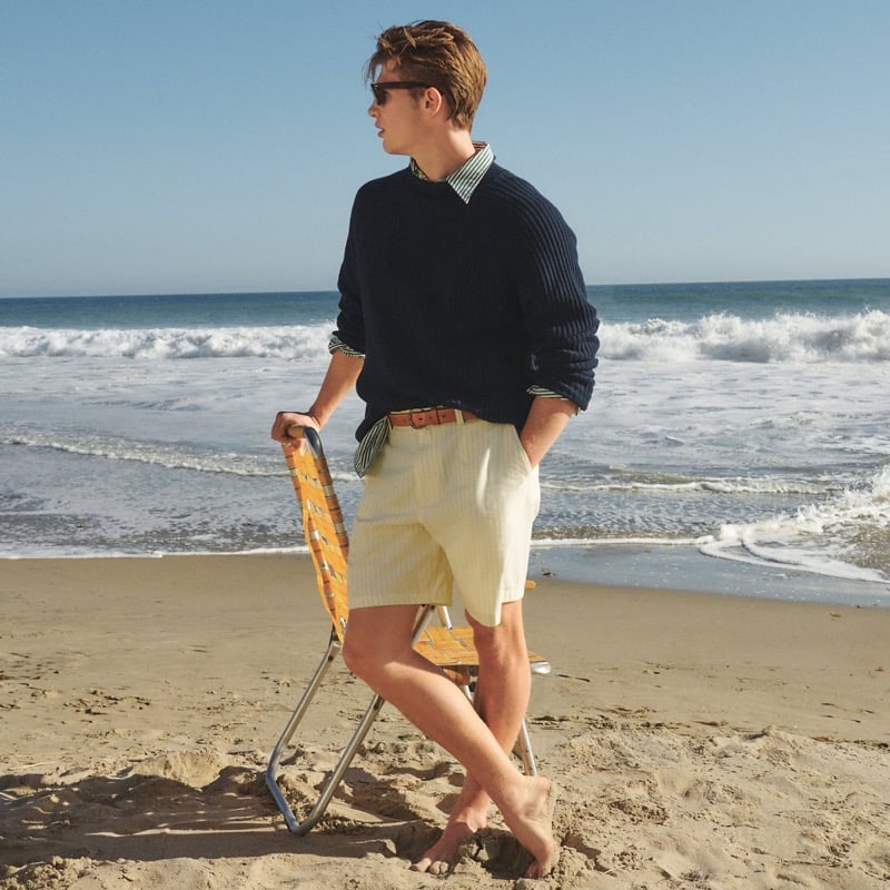 Joe stands barefoot on the beach in a J.Crew navy summer rollneck ribbed sweater layered over a green and white striped point-collar poplin shirt, with a brown leather belt and ivory and yellow striped classic chino shorts, holding a folded wooden beach chair