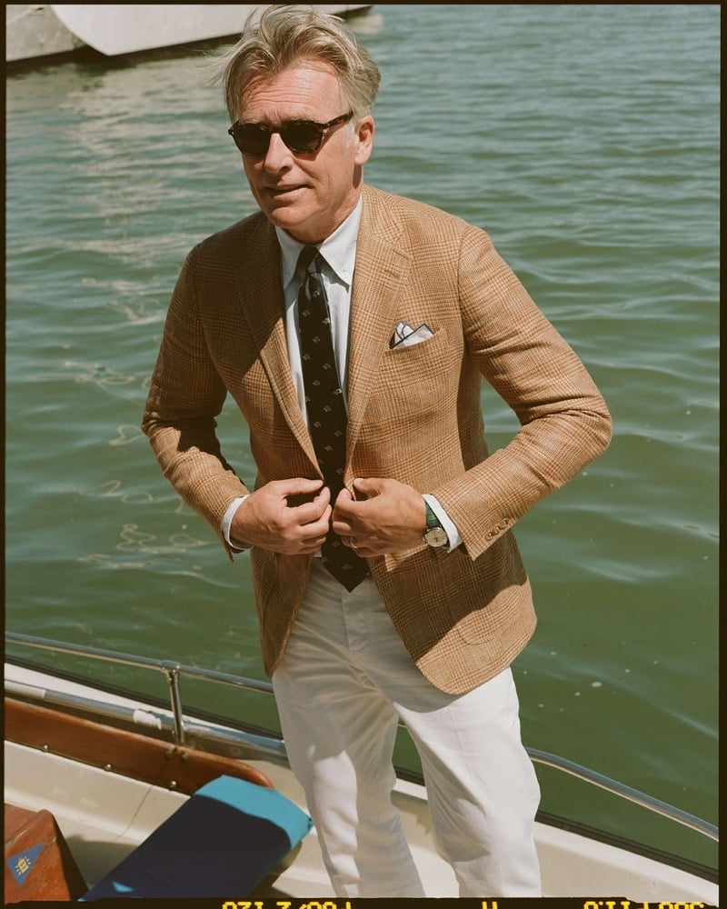 John Pearson wears tortoiseshell sunglasses and a rust glen check sport coat over a white shirt and navy patterned tie with white trousers, stepping onto a boat at the harbor