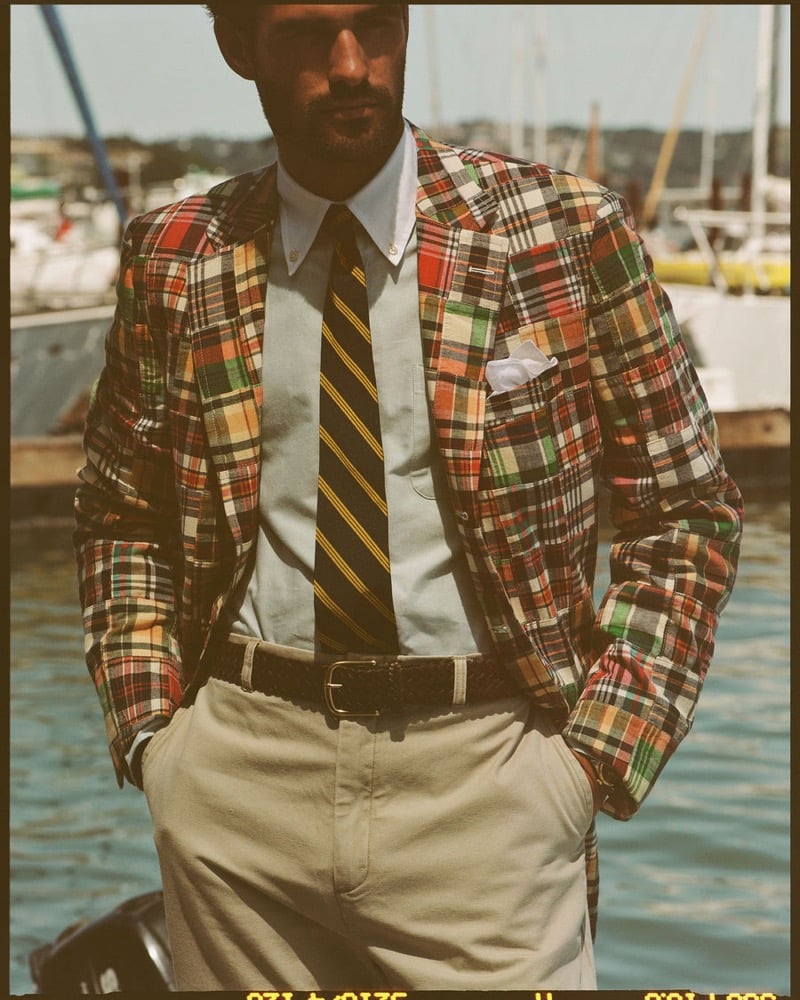 João Forte wears a patchwork madras sport coat over a pale blue shirt with a navy and gold striped tie, khaki trousers, and a brown woven leather belt at the marina