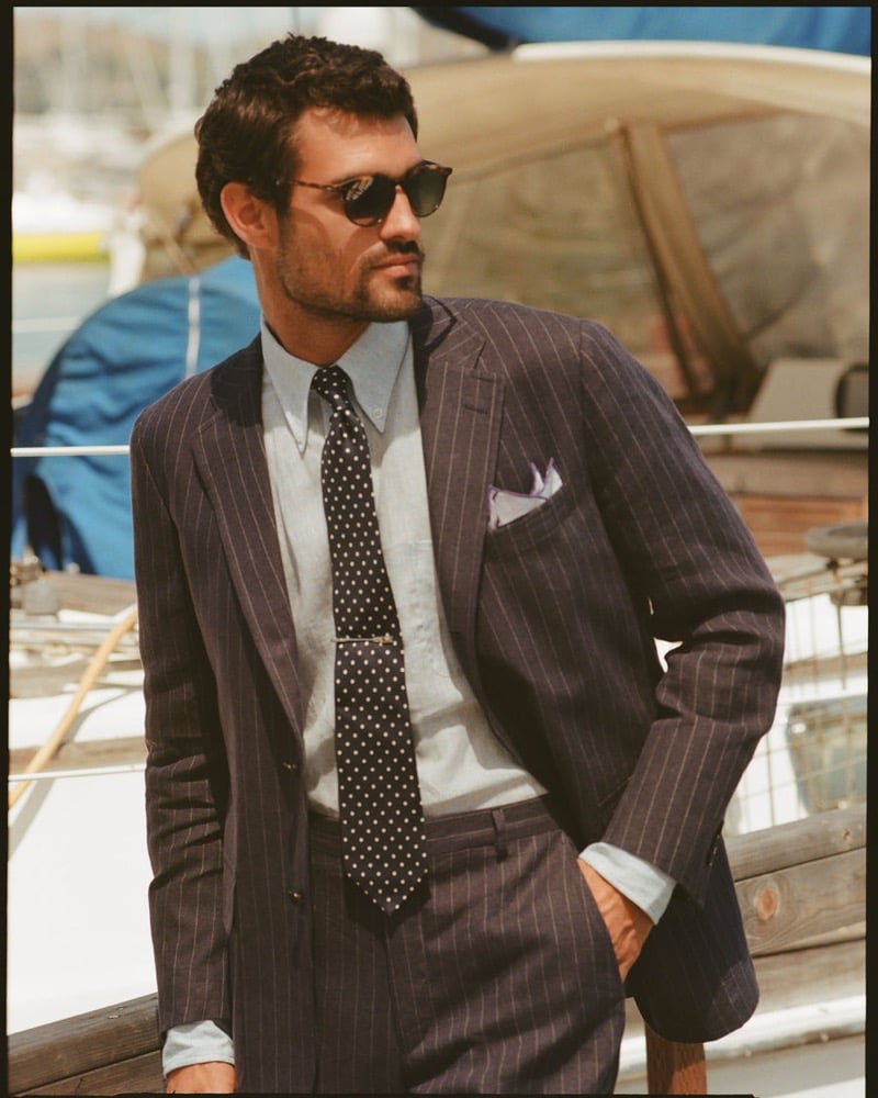 João Forte wears tortoiseshell sunglasses and a navy pinstripe suit over a chambray shirt with a navy polka dot tie and lavender pocket square aboard a sailboat