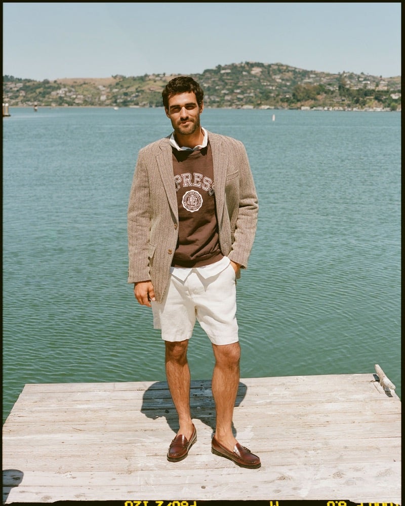João Forte wears a tan herringbone sport coat over a brown J. Press crest sweatshirt and white shirt with cream linen shorts and burgundy penny loafers on a wooden dock