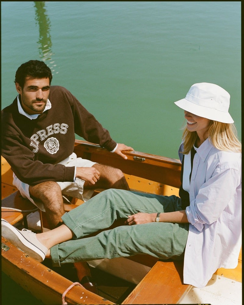 João Forte wears a brown J. Press crest sweatshirt with cream shorts and white canvas sneakers seated in a wooden rowboat beside a blonde woman in a white bucket hat, pale blue shirt, and green trousers