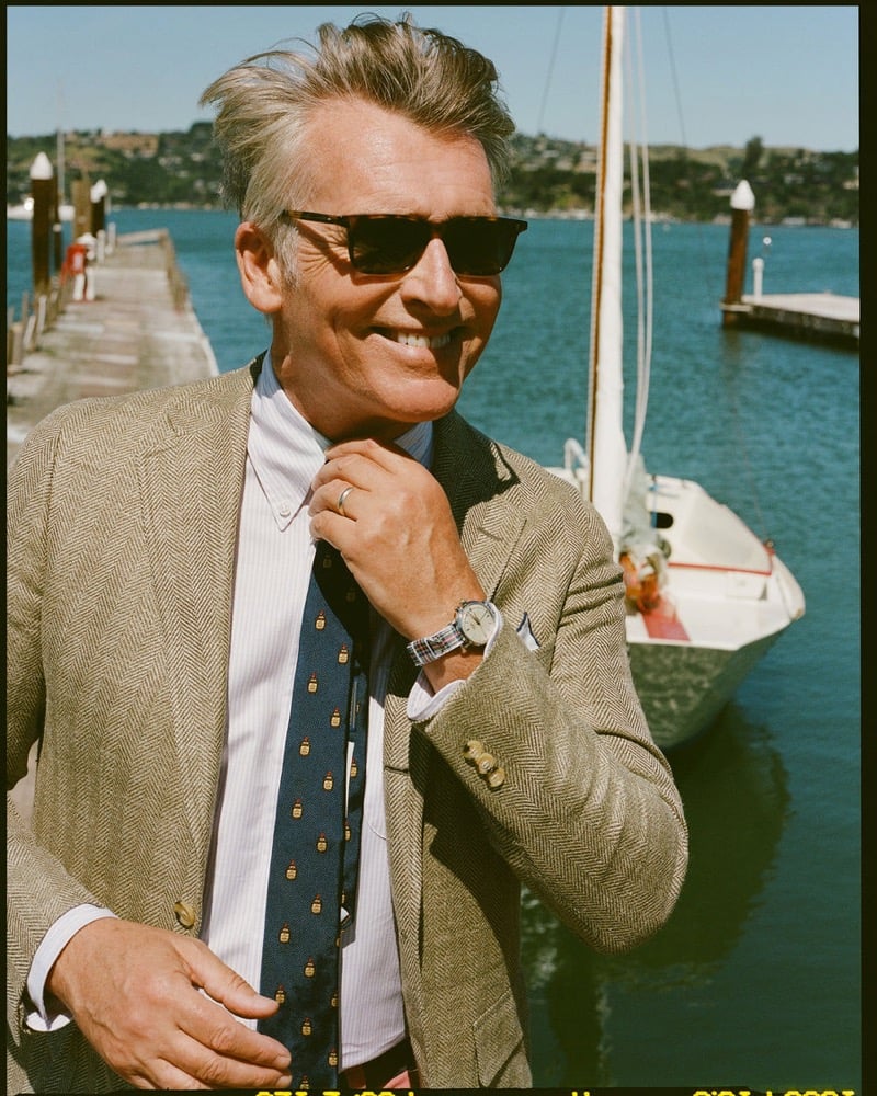 John Pearson wears tortoiseshell sunglasses and a tan herringbone sport coat over a pink and white striped shirt with a navy tie patterned with embroidered motifs at the marina