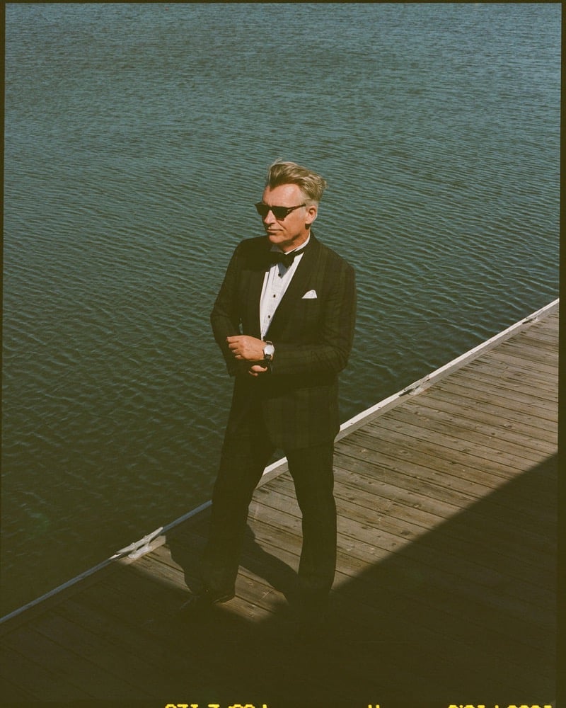 John Pearson wears tortoiseshell sunglasses and a black tuxedo with bow tie and white pocket square standing on a wooden dock above the water
