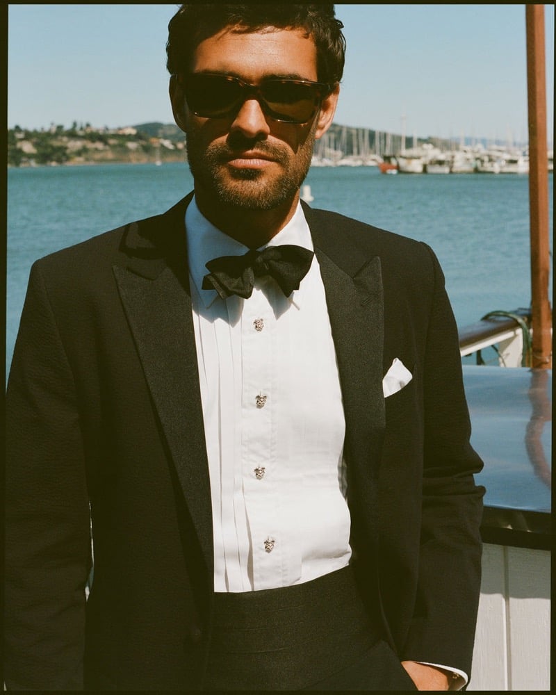 João Forte wears tortoiseshell sunglasses and a black tuxedo with a black bow tie over a pleated white shirt with crest buttons and a white pocket square on a yacht
