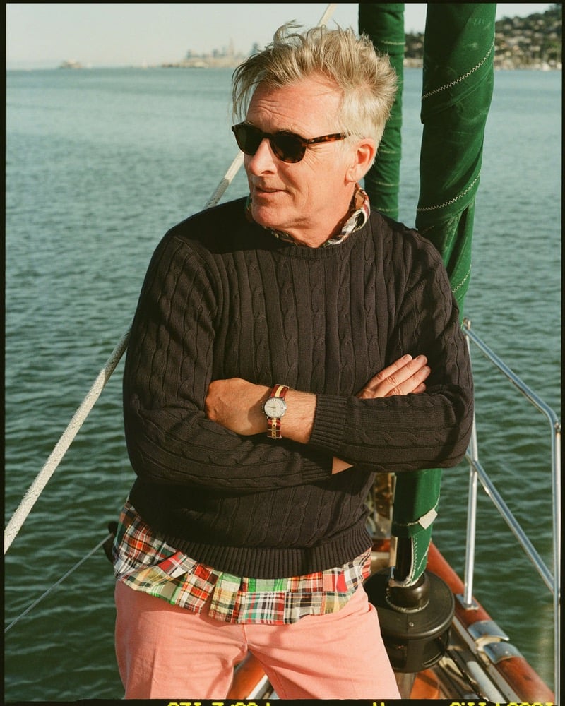 John Pearson wears tortoiseshell sunglasses and a navy cable-knit sweater over a patchwork madras button-down with salmon pink shorts aboard a sailboat