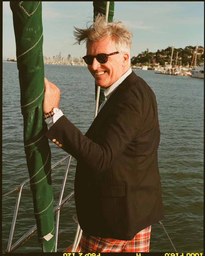 John Pearson wears tortoiseshell sunglasses and a navy blazer with gold buttons over a blue striped shirt and green knit tie with red plaid madras shorts on a sailboat
