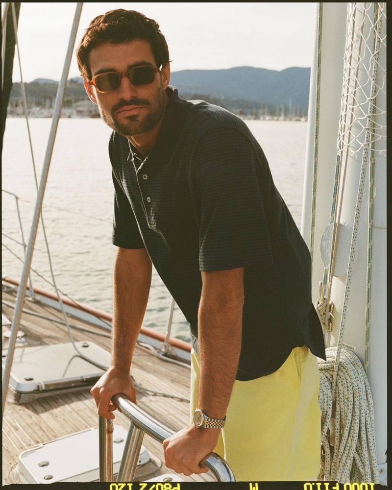João Forte wears tortoiseshell sunglasses and a navy striped short-sleeve polo with yellow trousers and a gold watch at the wheel of a sailboat