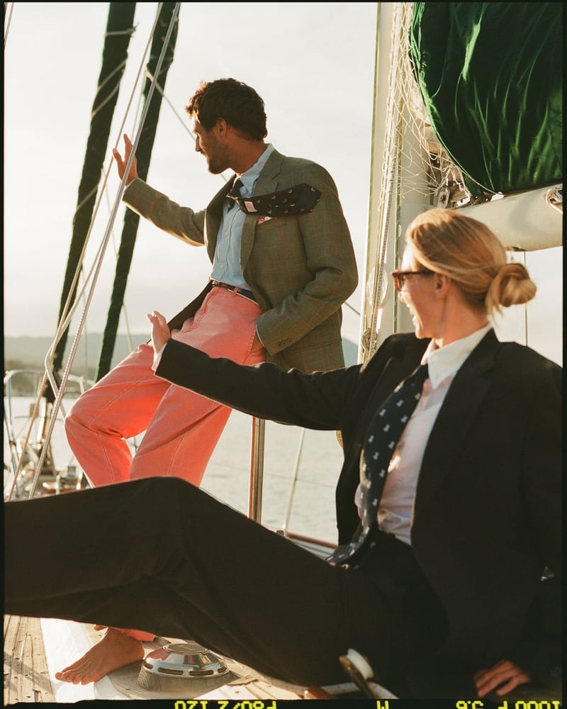 João Forte wears a green and grey windowpane sport coat over a chambray shirt with a navy floral tie and salmon trousers beside a blonde woman in a black blazer, white shirt, and navy patterned tie aboard a sailboat