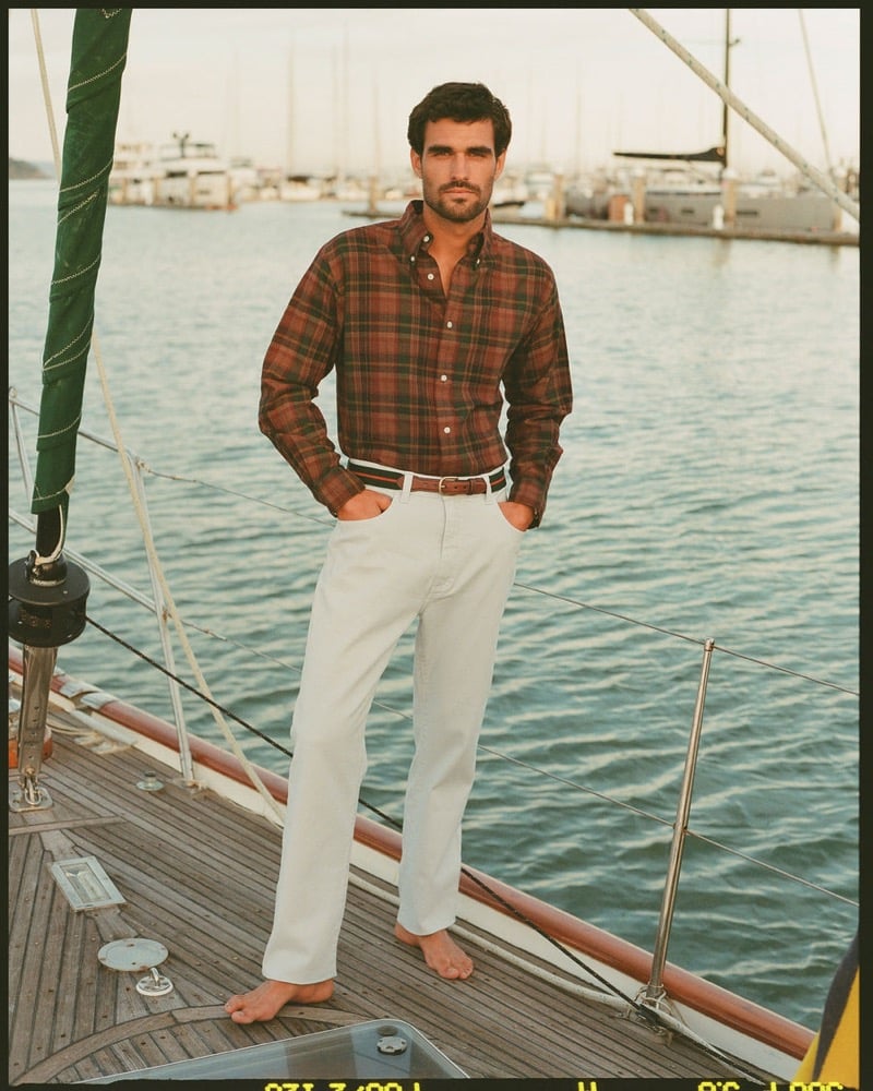 João Forte wears a red and black plaid flannel button-down with white jeans and a striped webbing belt standing barefoot on a sailboat deck