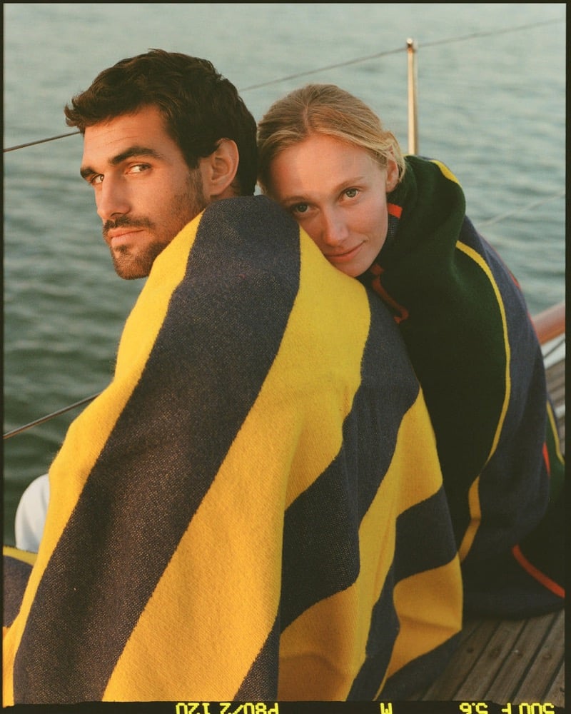 João Forte and a blonde woman wrap themselves in a navy and yellow striped wool blanket aboard a sailboat at sunset