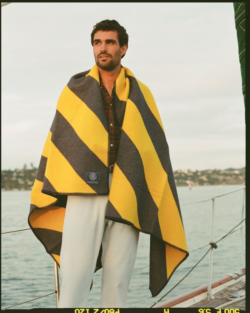 João Forte wraps himself in a navy and yellow striped Faribault Mill wool blanket over a brown plaid shirt with white trousers on a sailboat