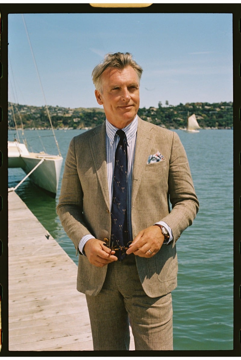 John Pearson wears a tan linen suit over a blue and white striped shirt with a navy patterned tie and floral pocket square, holding sunglasses on a dock beside a sailboat