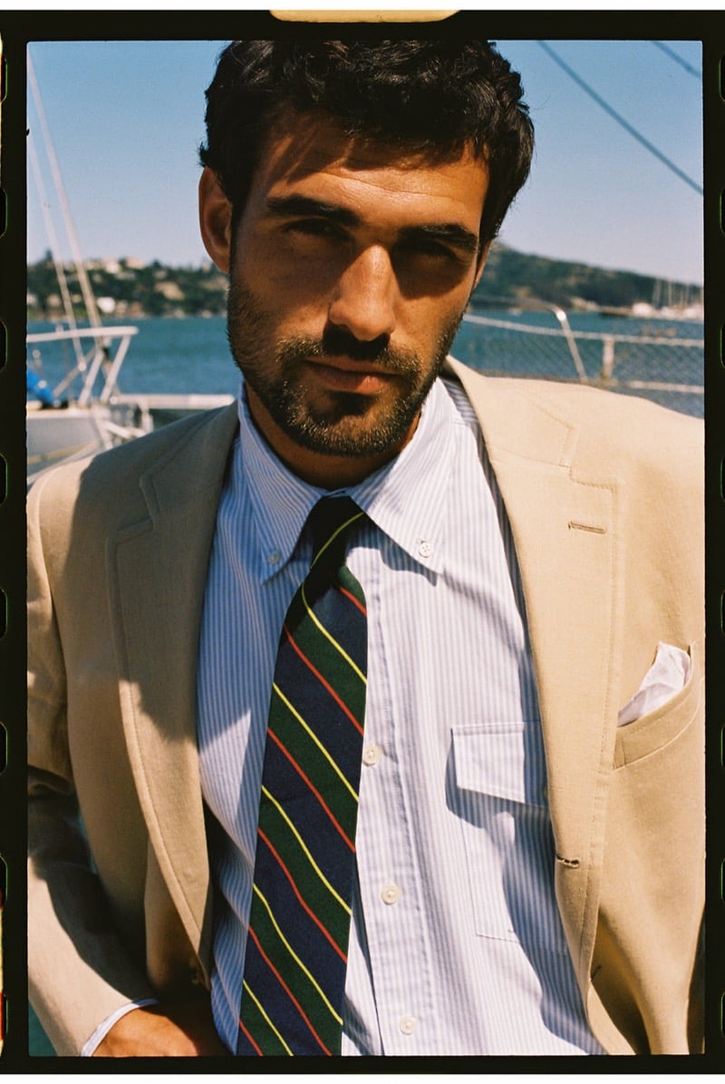 João Forte wears a tan linen sport coat over a blue and white striped shirt with a multicolor repp stripe tie and a white pocket square at the marina