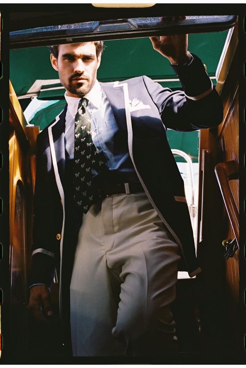 João Forte wears a navy blazer with white piping and gold buttons over a white shirt with a green embroidered motif tie and taupe trousers, stepping through a doorway