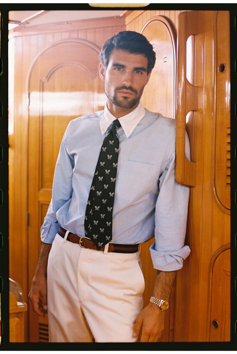 João Forte wears a pale blue button-down shirt with a black tie patterned with crossed tennis rackets, cream trousers, and a brown woven leather belt against a wood-panelled wall