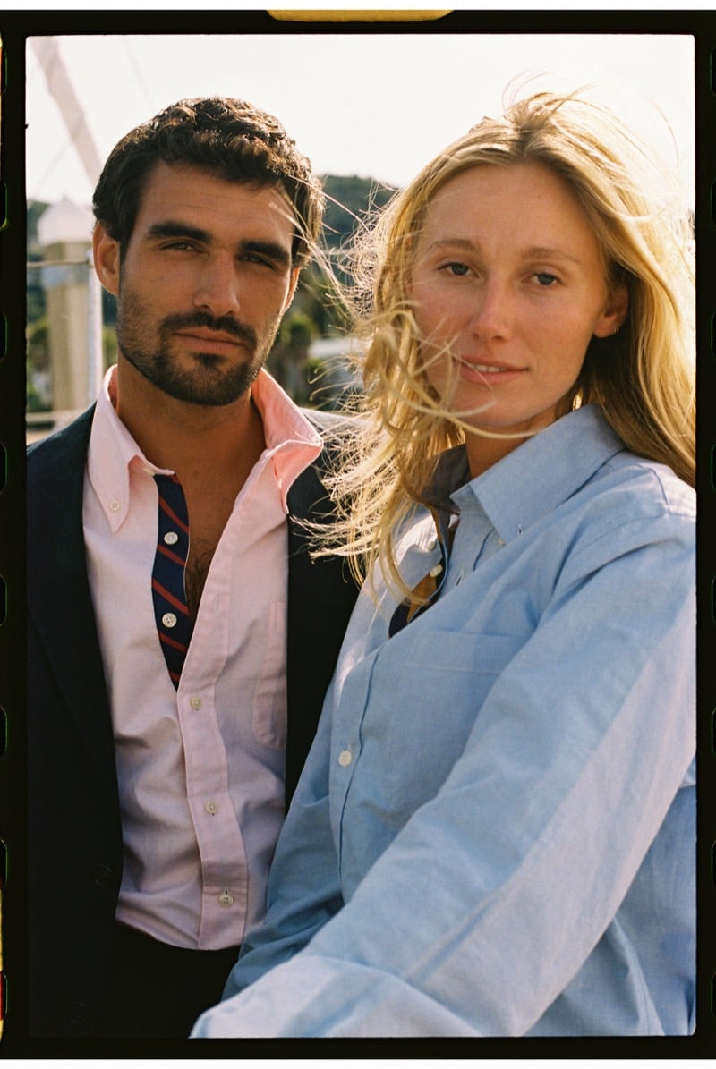 João Forte wears a navy blazer over a pink button-down shirt with a navy and red striped tie beside a blonde woman in a pale blue button-down shirt at the harbor