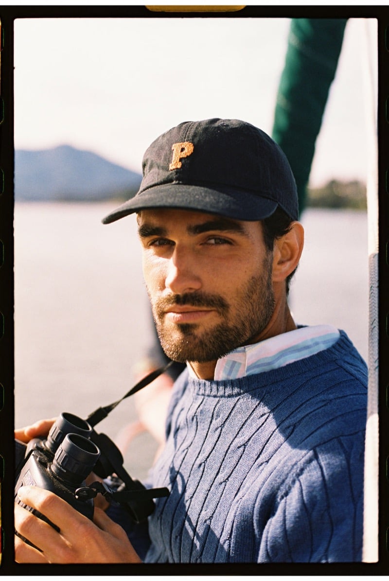 João Forte wears a navy baseball cap with an orange P emblem and a blue cable-knit sweater over a pink and blue striped button-down, holding a pair of black binoculars on a sailboat