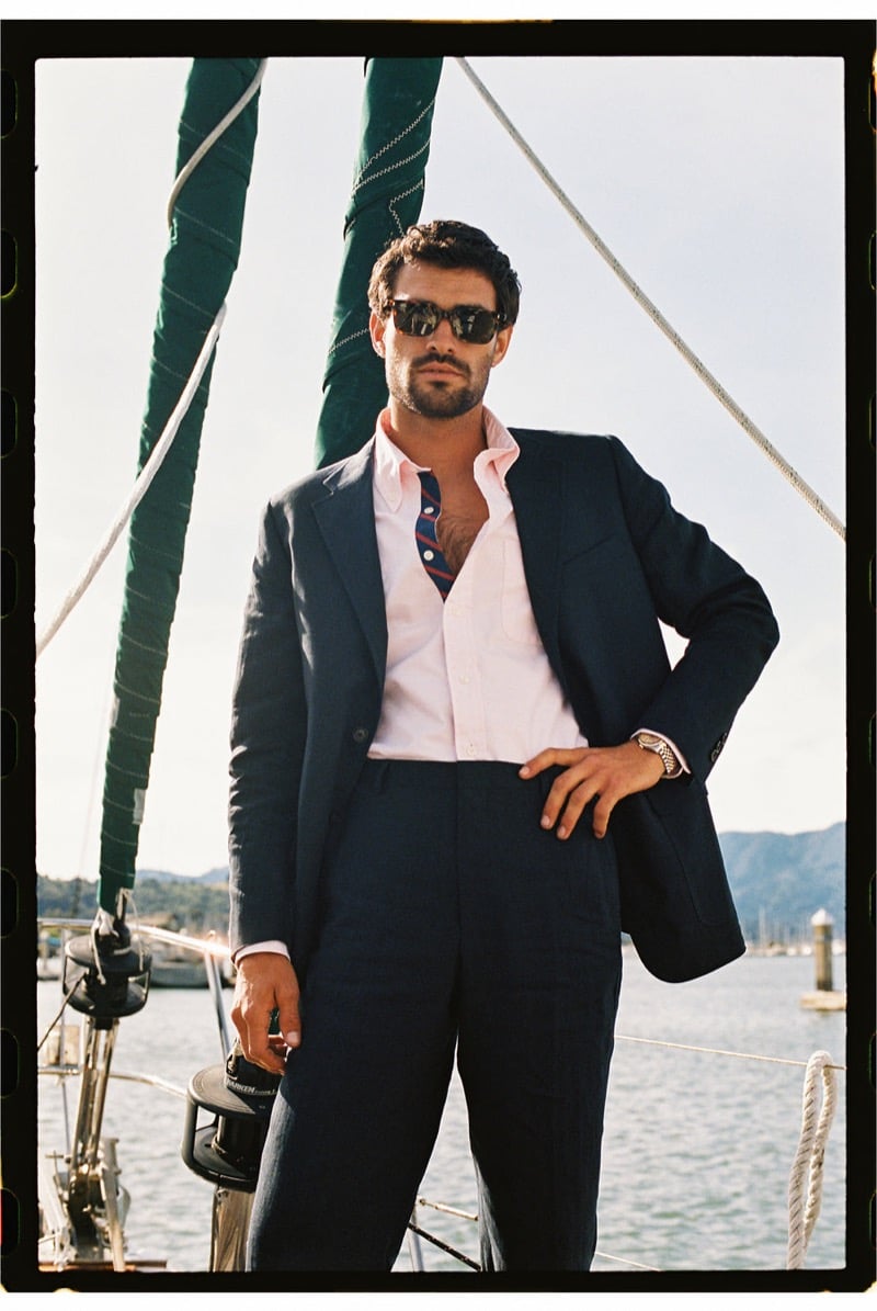 João Forte wears tortoiseshell sunglasses and a navy linen suit over an unbuttoned pink shirt with a loose navy and red striped tie aboard a sailboat