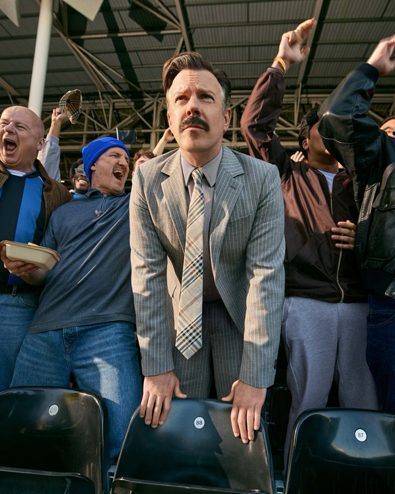Jason Sudeikis stands above empty stadium seats among cheering fans, wearing a gray pinstripe two-button suit, a white shirt, and a Burberry check tie