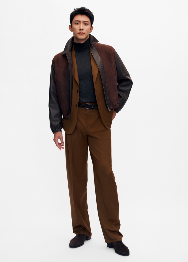 Johnny Huang stands against a pale backdrop in a dark leather bomber jacket with a brown suede yoke, layered over a brown wool blazer, dark gray turtleneck, brown pleated wool trousers, and dark suede shoes