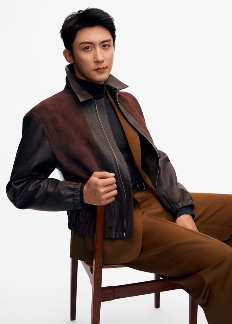 Johnny Huang sits on a wood chair in a dark leather bomber jacket with a brown suede yoke over a brown wool jacket, a dark turtleneck, and pleated brown trousers