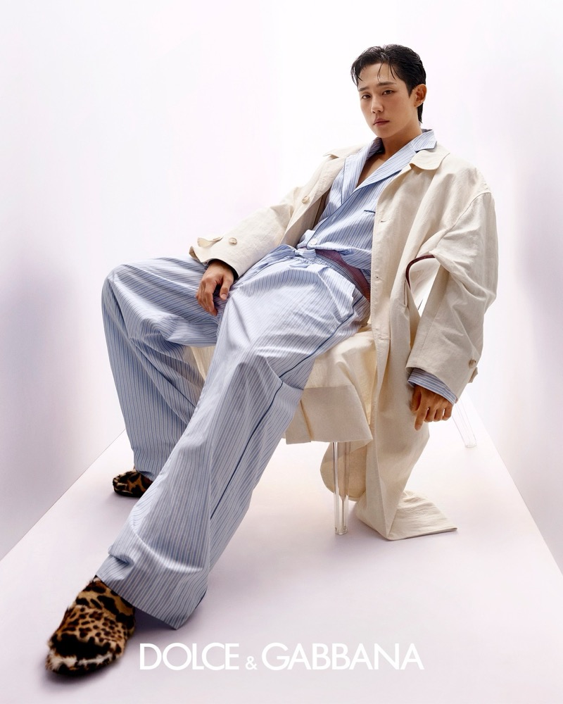 Jung sits sideways on a transparent acrylic chair in a sky blue pinstripe pajama-style shirt and matching wide-leg trousers with an ecru trench coat and leopard print slippers on his feet