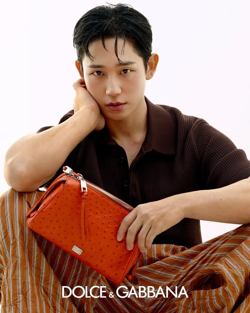 Jung rests his chin on one hand in a dark chocolate ribbed knit short-sleeve polo, cradling an orange ostrich leather zip pouch with silver hardware against his lap, a striped trouser visible beneath