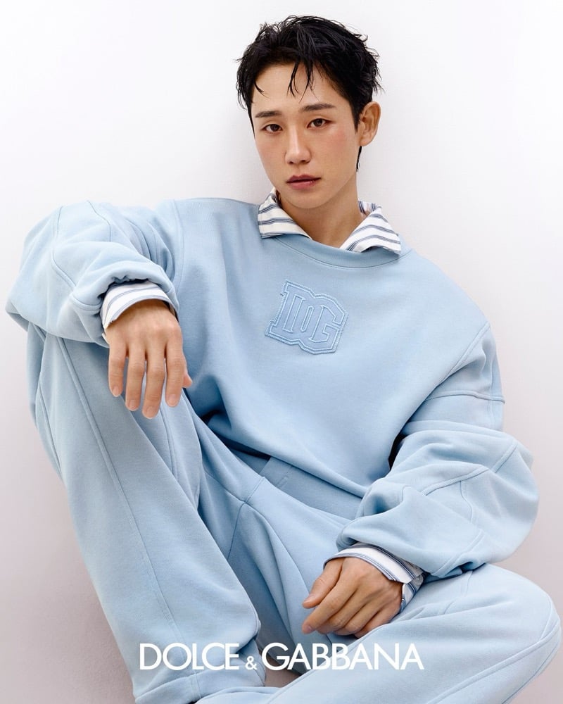 Jung sits with one knee raised in a pale powder blue oversized sweatshirt with a tonal DG monogram patch at the chest, layered over a navy and white pinstripe shirt visible at the collar and cuffs, paired with matching powder blue sweatpants
