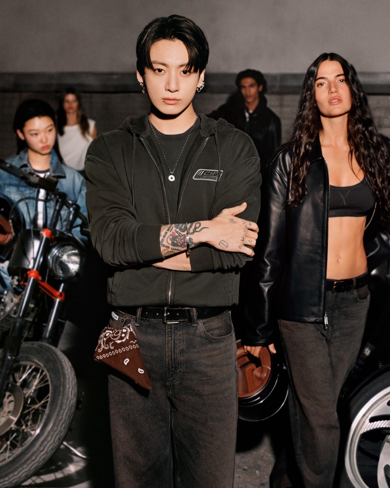 Jung Kook stands at center in a garage of vintage motorcycles in a washed dark green zip hoodie over a CKJK logo tee, dark wide-leg jeans, and a brown bandana tied at the hip, models surrounding him