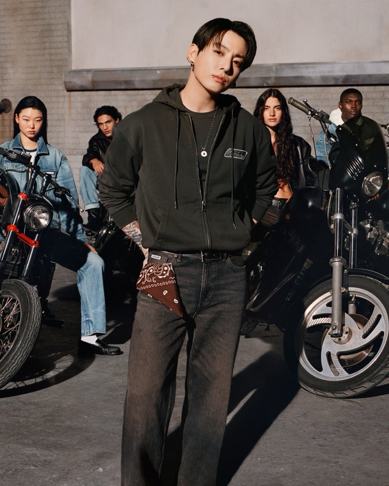 Jung Kook stands in a motorcycle garage in a washed dark green zip-up hoodie over a black CKJK logo tee, dark jeans, and a brown paisley bandana tied at his waist