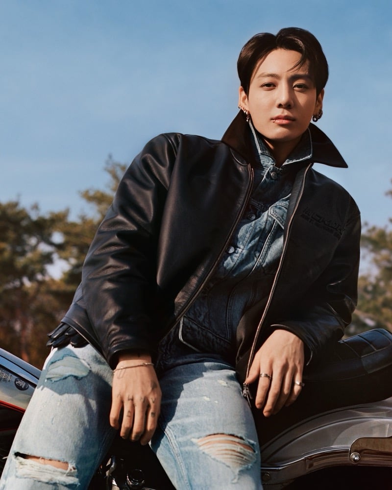 Jung Kook sits on a motorcycle in a black leather biker jacket layered over a denim trucker jacket, paired with light-wash ripped straight jeans against a blue sky and bare trees