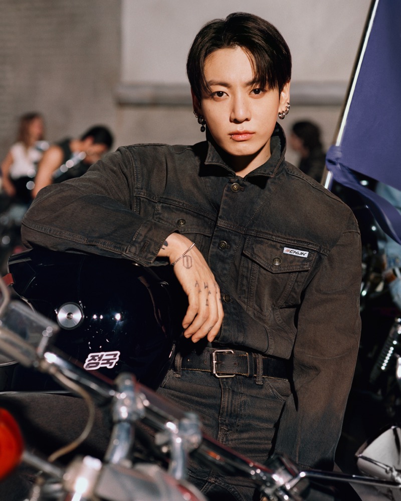 Jung Kook leans on a black motorcycle helmet stamped with the Korean characters for his name, dressed in a black-washed denim work shirt with snap chest pockets and a black belt