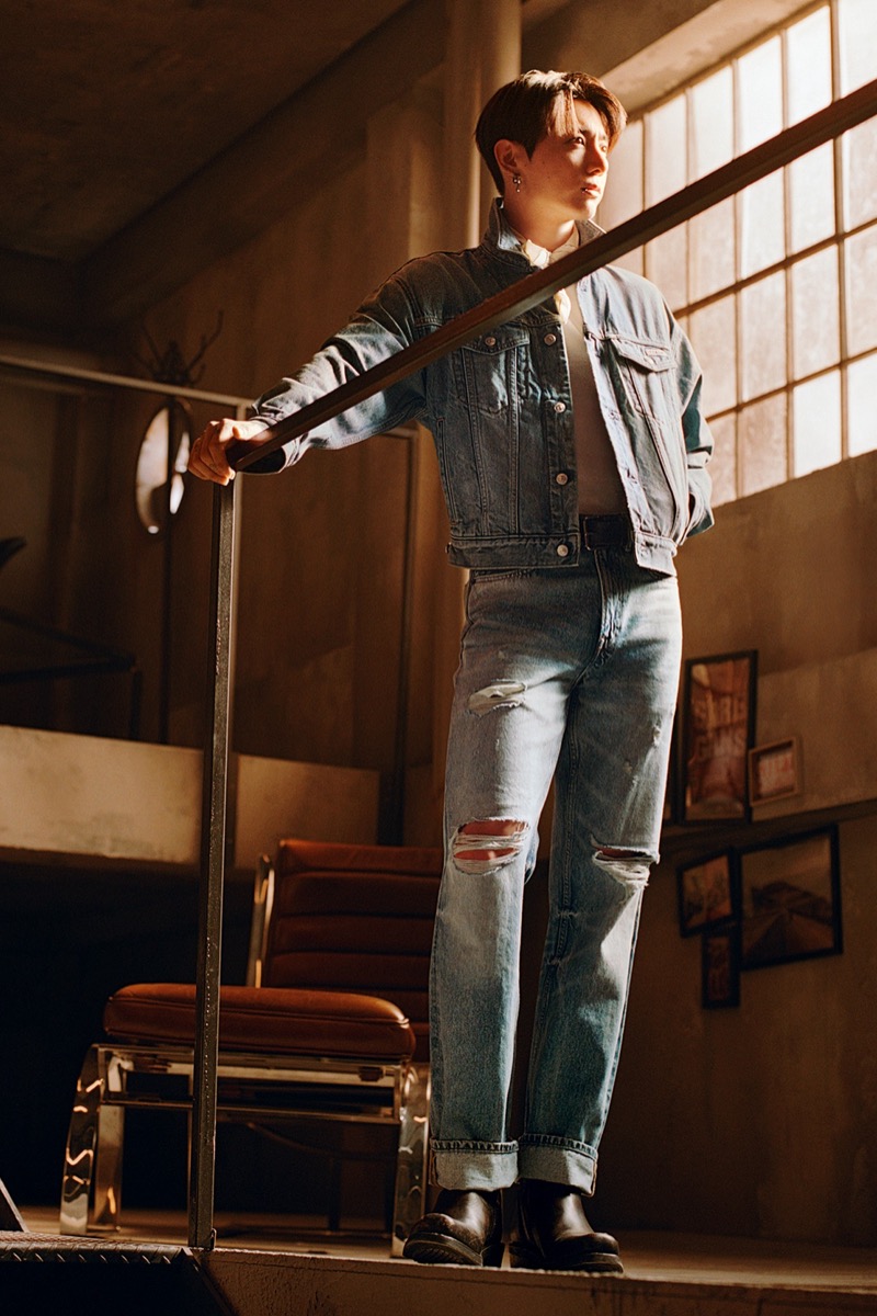 Jung Kook stands on a raised concrete platform inside an industrial loft in a cropped light-wash denim trucker jacket over a white tee, light-wash straight-leg ripped jeans, and black leather boots, tall industrial windows behind him