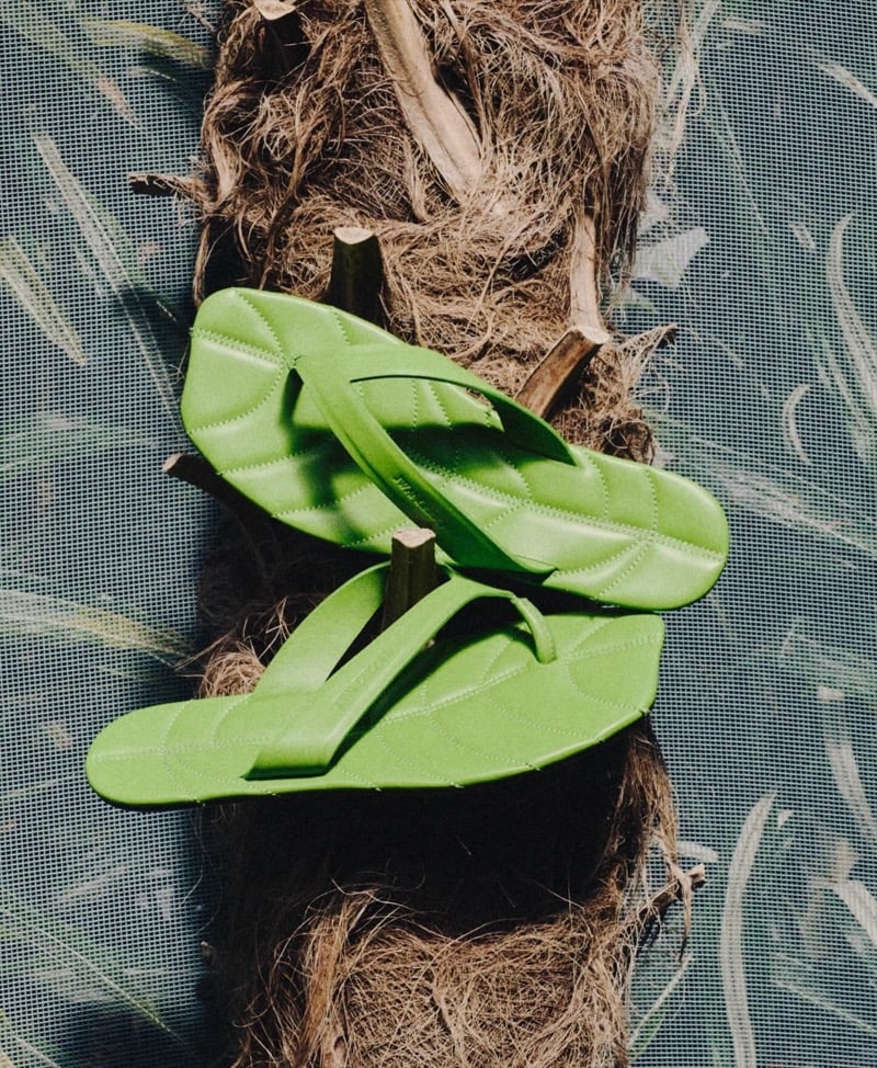 A pair of green leather thong slides shaped like overlapping leaves rest on a coconut fiber surface