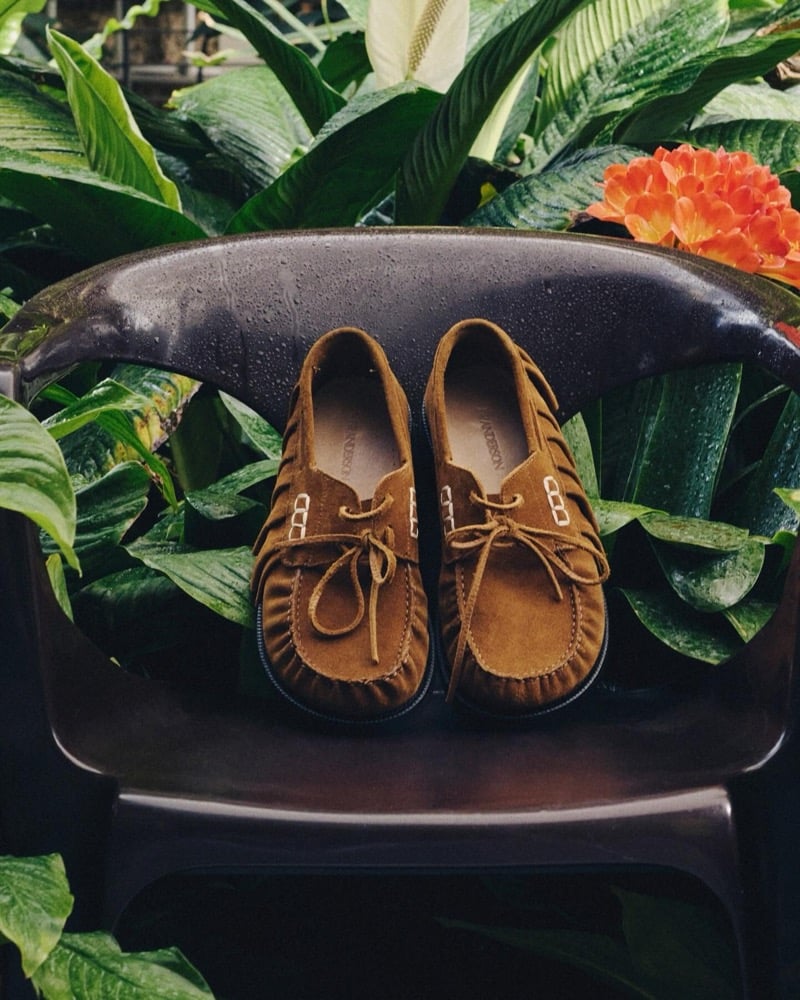 A pair of tan suede ruffled boat shoes sits on a glossy black chair surrounded by tropical leaves and an orange clivia bloom