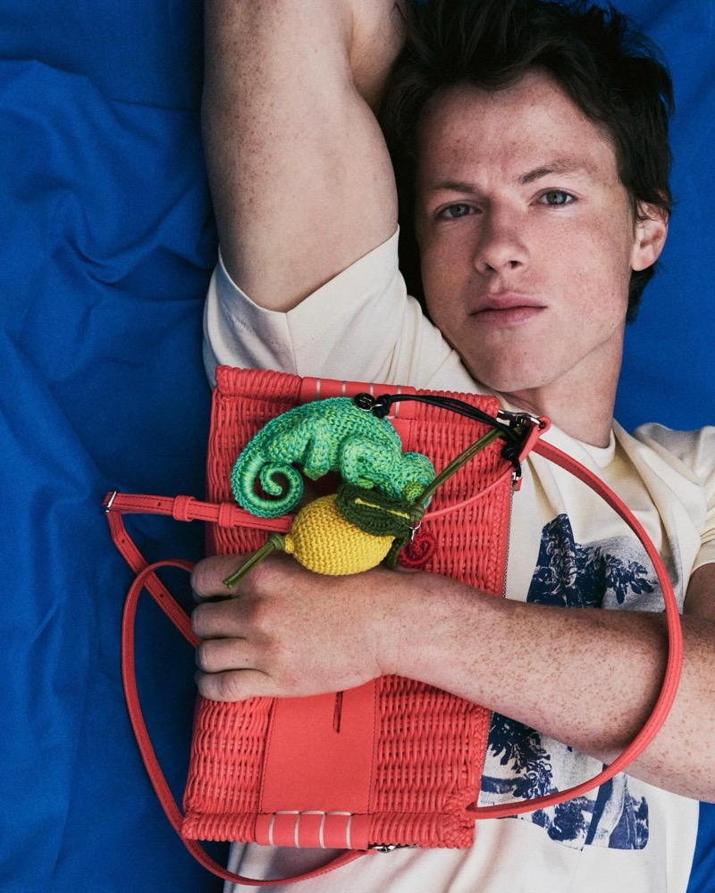 Milo lies on a cobalt blue sheet holding a coral woven leather tote bag with a green crochet chameleon and yellow crochet lemon charm, wearing a cream t-shirt with a printed graphic
